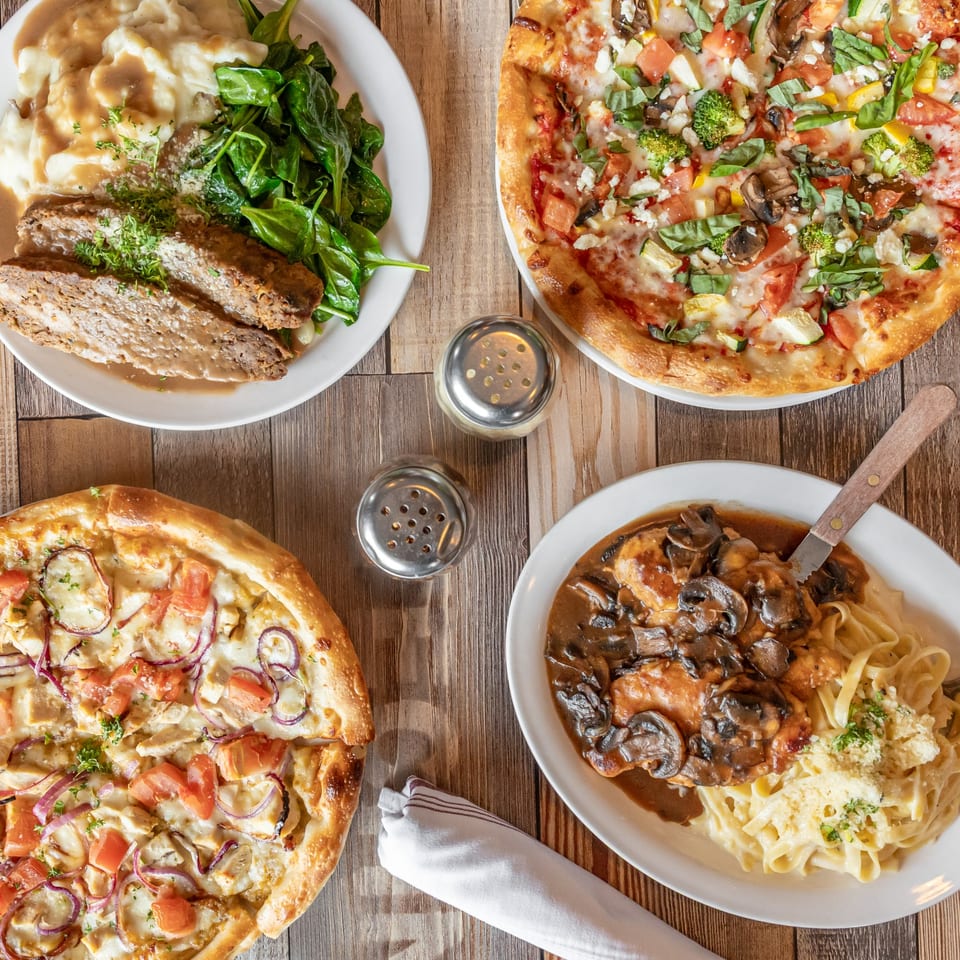 Overhead view of a wooden table with meatloaf, two pizzas, and chicken marsala with pasta.