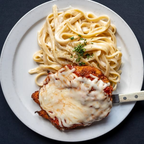 Chicken parmesan with melted mozzarella and marinara sauce, served with fettuccine alfredo on a white plate.
