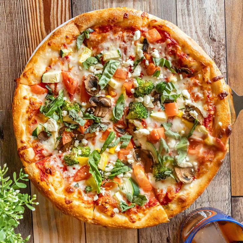 A vibrant vegetarian pizza with broccoli, mushrooms, tomatoes, zucchini, and feta cheese on a wooden table.