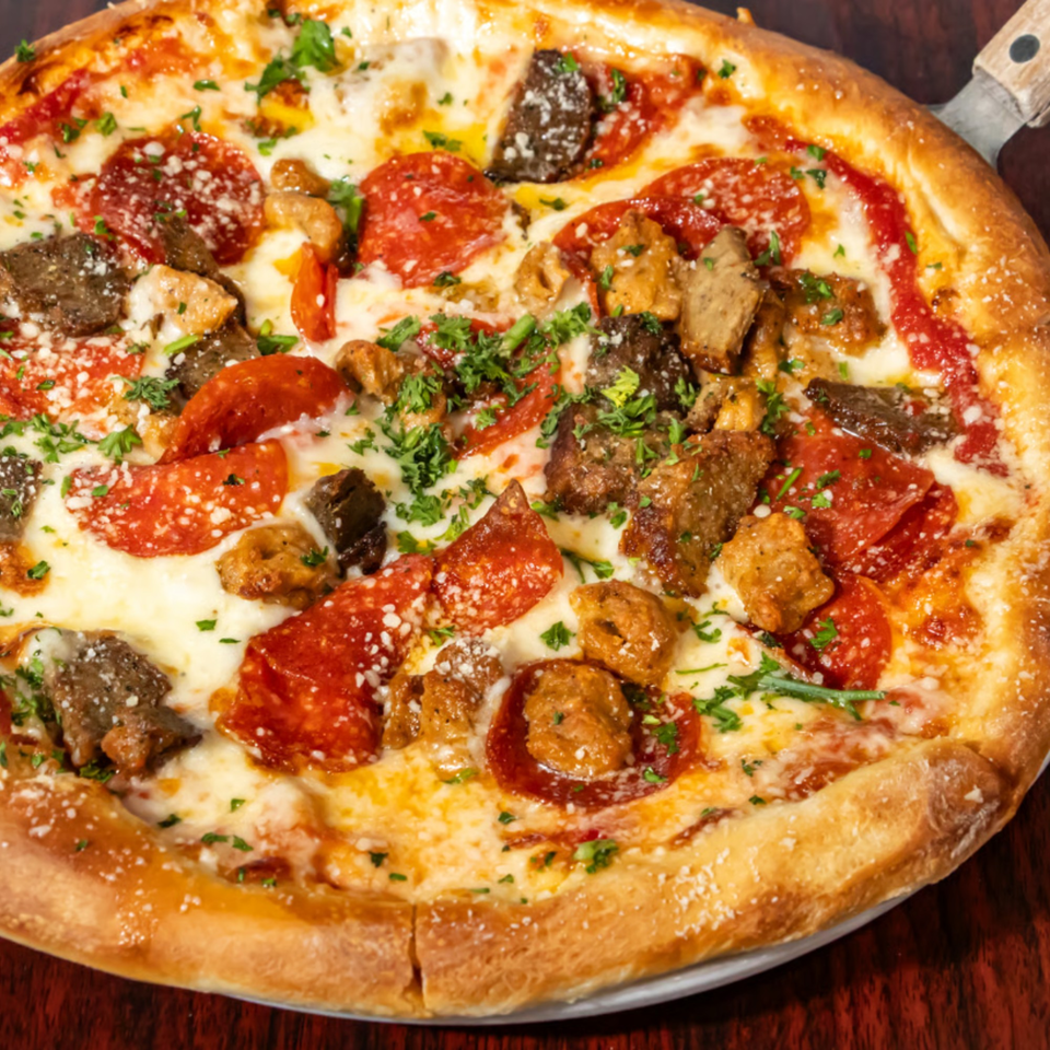 Close-up of a pizza with pepperoni, sausage, steak, and fresh parsley on a wooden table.