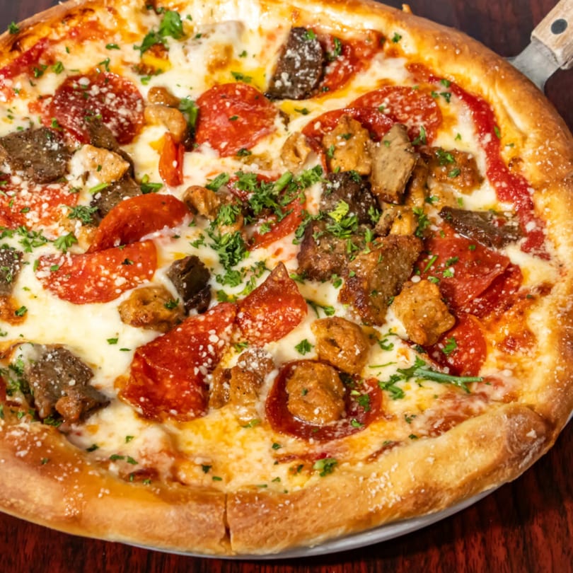 Close-up of a pizza with pepperoni, sausage, steak, and fresh parsley on a wooden table.