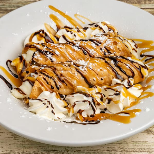 Three cannoli drizzled with chocolate and caramel, topped with whipped cream and powdered sugar.