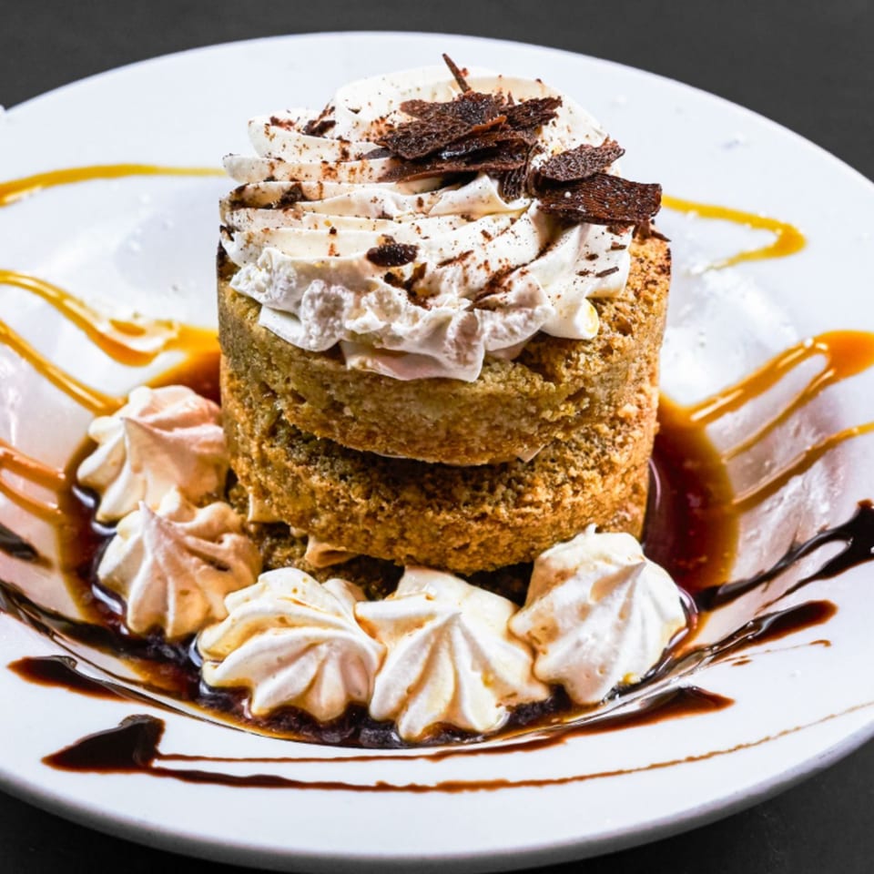Layered dessert with whipped cream, chocolate shavings, and meringue, drizzled with caramel and chocolate sauce.