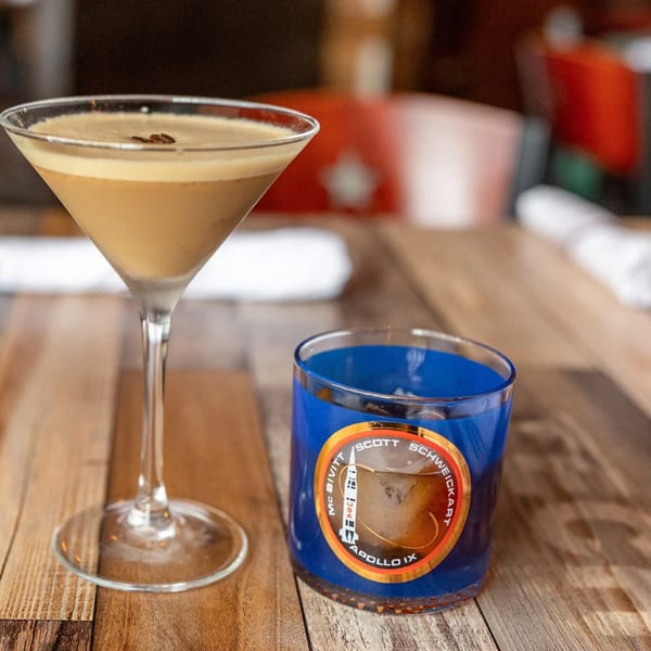 Espresso martini with coffee beans and a blue glass with an Apollo IX logo on a wooden table.