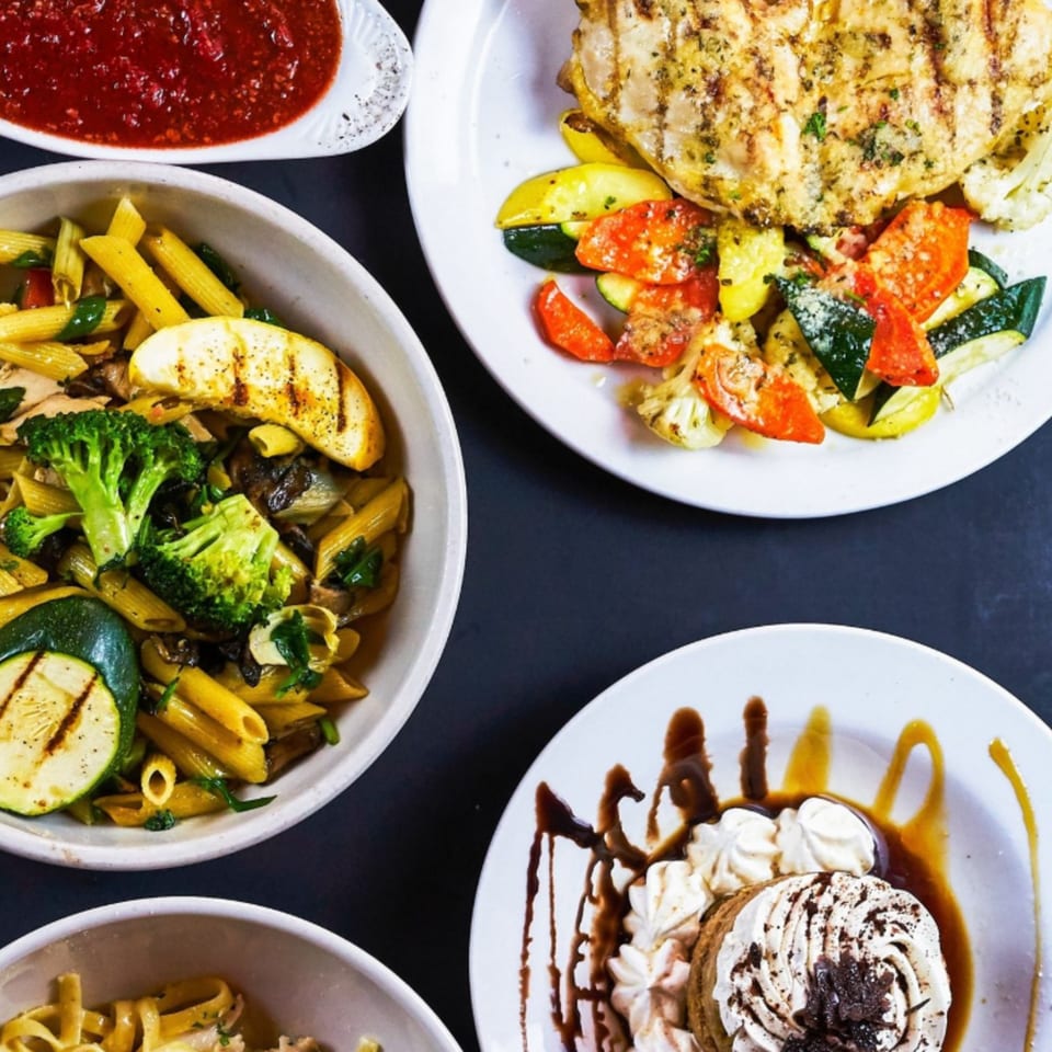 Overhead view of grilled chicken with vegetables, pasta with broccoli, and a dessert on a dark surface.