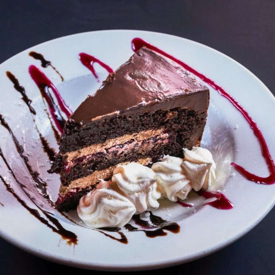 A slice of chocolate cake with whipped cream and berry and chocolate drizzles on a white plate.