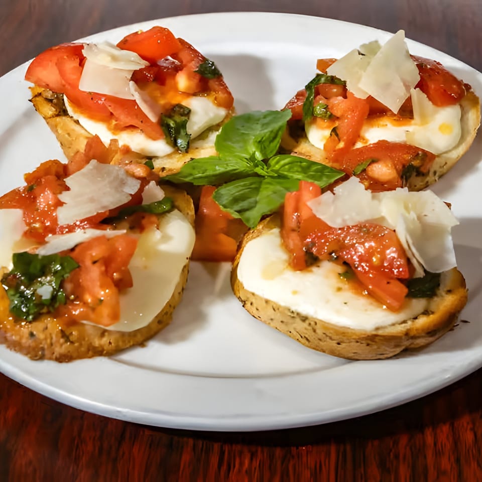 Four bruschetta appetizers with tomatoes, mozzarella, basil, and parmesan on a white plate.